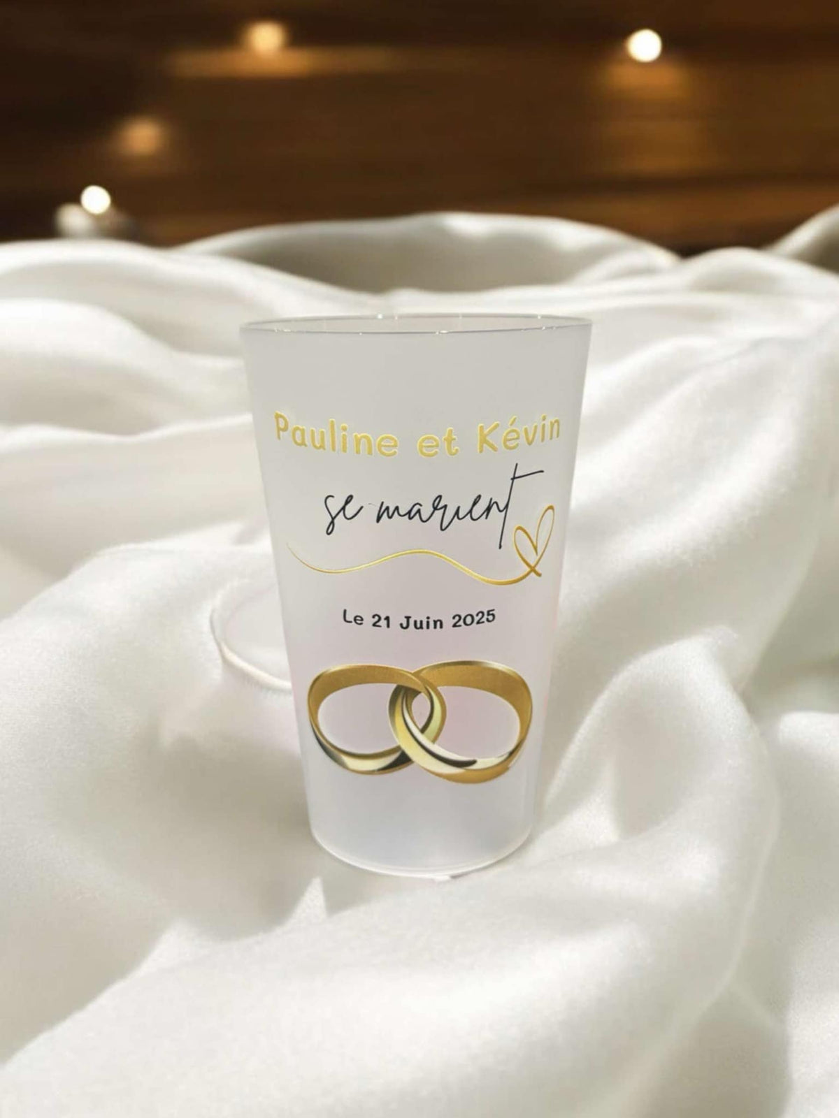 Gobelet de mariage personnalisé au texte doré, posé sur du satin blanc à côté des alliances